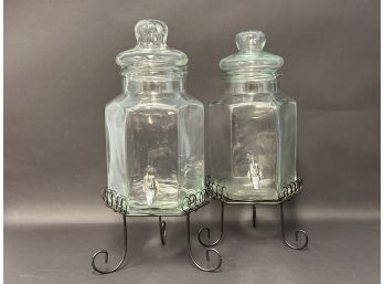 A Pair Of Glass Beverage Dispensers With Stands